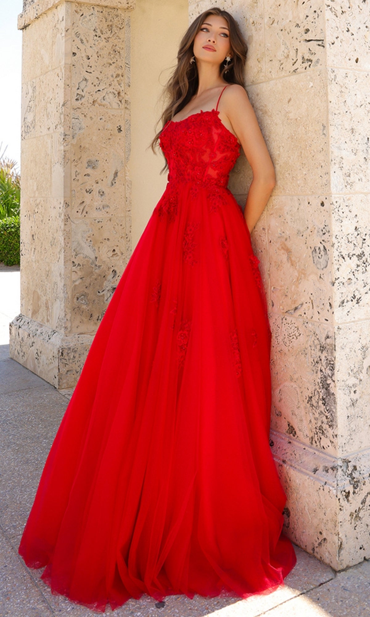 A woman stands outdoors against a textured stone wall wearing the Embroidered-Tulle Long A-Line Prom Ball Gown 7035, a sleeveless, floor-length red dress with delicate lace and floral embellishments.